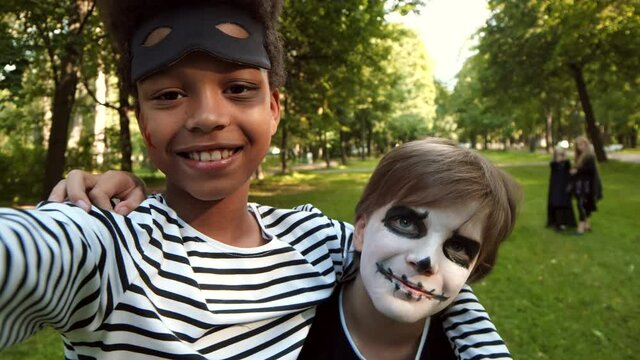 Close up of two diverse boys wearing costumes of robber and skeleton are standing together in the park and making selfies