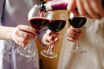 Pour red wine. Bride and bridesmaids toasting with champagne and having fun on wedding morning. Hands holding glasses. Close up photo of girls celebrating a bachelorette party.