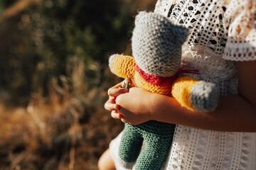 Happy girl playing with a knitted toy  in nature during summer time. Girl in a white dress with toy in hands on field.