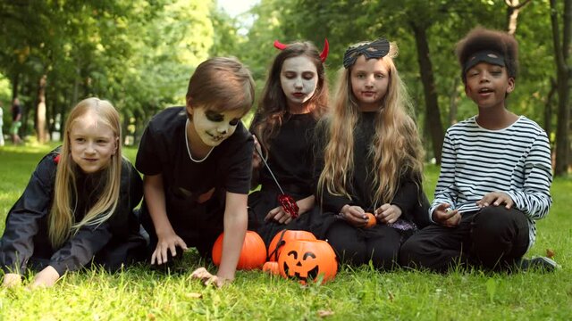 Wide shot of three Caucasian girls and two multiethnic boys wearing Halloween costumes are sitting on grass outdoors, eating sweets and scaring with their hands and fingers