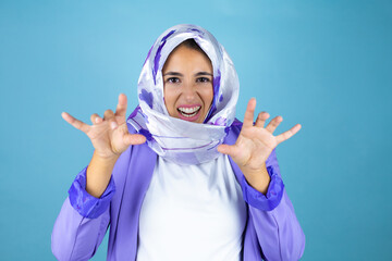 Young beautiful arab woman wearing islamic hijab over isolated blue background smiling funny doing...