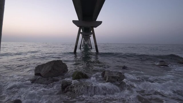 4K stock video dolly in and out among bridge pilars going into the sea. Waves breaking over rocks under a concrete structure at the beach. Ocean landscape at sunrise. Badalona old landmark