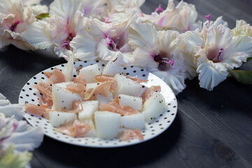 Plate with melon and prosciutto on black wooden background and white gladiolus flowers