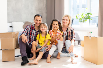 Fototapeta na wymiar Caucasian family, man, woman and two girls sit on floor, unpack boxes and smile in new house. Behind them moving boxes.