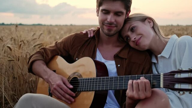 A young happy couple in love man and woman are sitting and hugging together while playing the guitar in the field