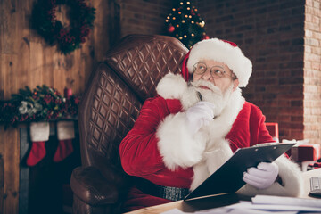 Minded pensive grey hair santa claus sit table prepare x-mas christmas event think thoughts write clipboard order wish list letters wear cap headwear in newyear advent spirit house indoors
