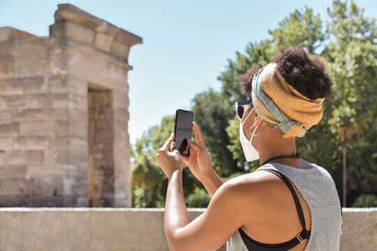 A young african origin woman wearing a face mask she uses her mobile phone to take a picture during a tourist excursion, on a blurred monumental background. Safe tourism in pandemic times.