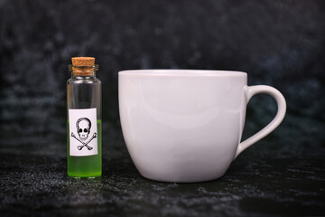 Small vial with cork and green liquid and poison skull label next to tea cup on dark background