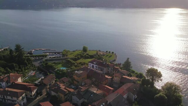 Magnificent sunset flight above Il sole di Ranco restaurant resort by Lake Maggiore and red terracotta village roofs with bright white shiny sun beam on water, Italy, overhead aerial circle approach 