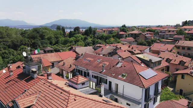 Scenic backward flyover above Il sole di Ranco restaurant, resort, and red terracotta village roofs with scenic view of Lake Maggiore mountain range in background, Italy, overhead aerial pull back