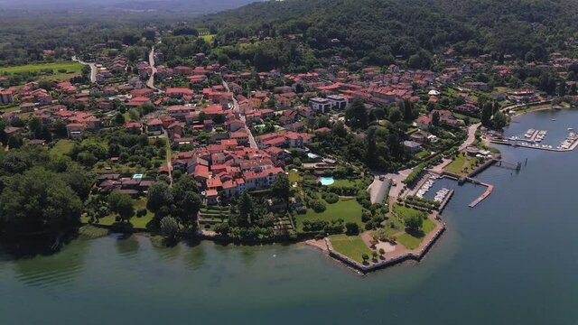 Establishing view of Il sole di Ranco restaurant and Lake Maggiore lakeside resort with red terracotta roofs, boat slips and mountain range in background, Italy, overhead aerial approach tilt down