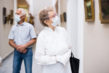 elderly European woman  in mask protecting against covid examines paintings on display in hall of...