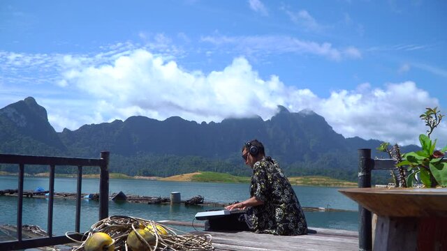 A man is sitting on the pier, playing a synthesizer, wearing black headphones, Cheo LAN, Kao SOK. view of beautiful limestone cliffs in a huge tropical lake surrounded by lush, dense rainforest 4K