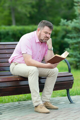 Obraz premium An adult man with short beard in summer clothes is reading a book while sitting on a bench in the park,