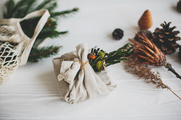 Stylish christmas gift wrapped in linen fabric, decorated with natural green branch on white rustic table background with pine cones and herbs. Zero waste winter holidays.