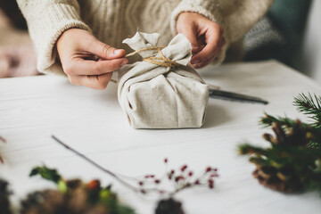 Female in cozy sweater preparing plastic free christmas present, zero waste. Hands wrapping stylish christmas gift in linen fabric on white rustic table with fir, pine cones, scissors, twine.