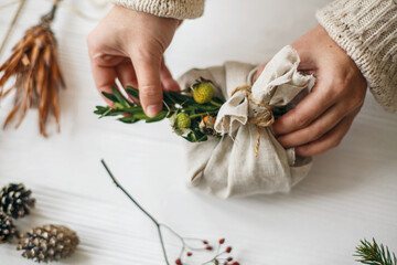 Hands decorating stylish christmas gift in linen fabric with green branch on white rustic table with natural herbs and pine cones. Female preparing plastic free christmas present. © sonyachny