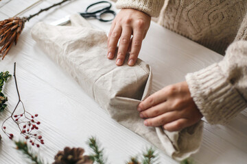 Plastic free christmas present, zero waste  holidays. Female hands in cozy sweater wrapping christmas gift in linen fabric on wooden table with green branch, pine cones, scissors.
