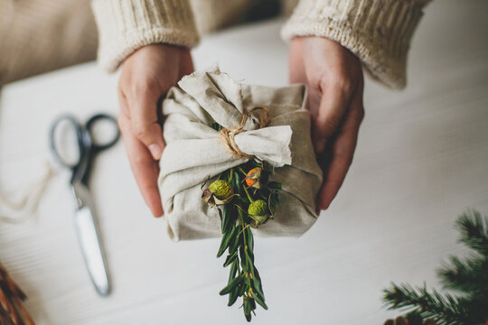 Zero waste Christmas gift. Hands holding stylish present wrapped in linen fabric with natural green branch on rustic background  with pine cones and scissors. Plastic free holidays
