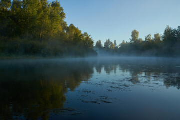 Fototapeta premium Autumn time. Dawn over the river in a misty, brooding haze. Beautiful view of the forest and river, covered with fog in the early morning. The sun's rays of light. September.