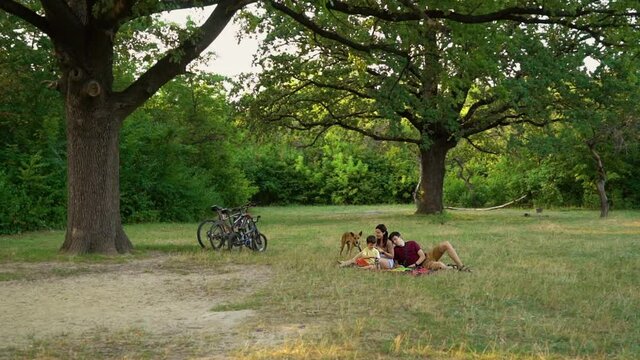 Happy family with dog relaxing and having picnic in green forest after biking. Arc shot caring owners feeding pet. Concept of food