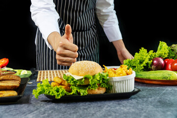 Hand of chef man thumb up with vegetable hamburger on table over black background. Homemade bread burger concept. Select focus