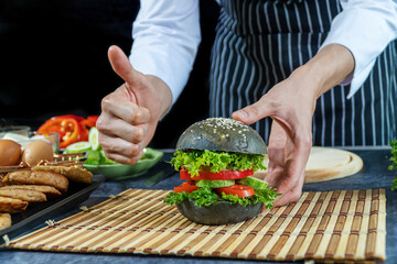 Hand of chef man thumb up with set vegetable hamburger on table over black background. Homemade bread burger concept.