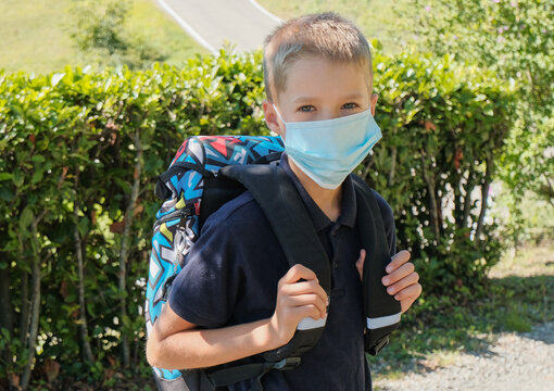 Caucasian blonde boy with a medical face mask close-up with backpack across green bushes on the backyard. School year during a pandemic. Education.
