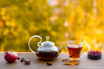 Autumn tea. Warming herbal tea in a glass teapot and cup on a wooden table against the backdrop of yellow trees. 
