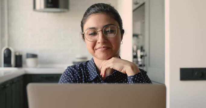Indian ethnicity 30s pretty business woman freelancer wearing eyeglasses do remote work telework distantly sit alone at desk using laptop portable device smiling enjoy fruitful workday at home office