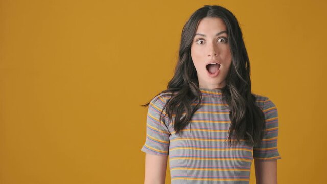 A shocked emotional young woman is pointing to the free space standing isolated over yellow background
