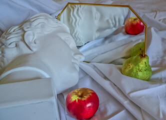 Ancient greek white sculpture plaster copy marble head of diana reflected in a mirror in gold frame against a background of white drapery fabric with folds with green pear and red yellow apple