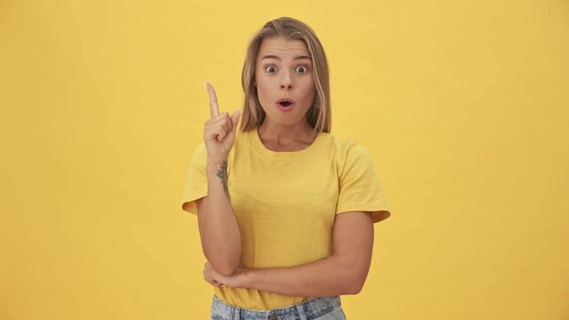 A happy young blonde woman in yellow T-shirt is thinking about something and come-up with an idea while raising her finger standing isolated over yellow background