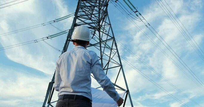 A man in a white protective helmet stands facing a high-voltage power line pole with a paper drawing unfolded in his hands, turns towards the camera.