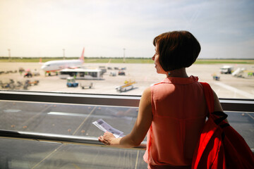Brunette caucasian woman indoors airport in gate and looking through the window on the aircrafts  preparing for departure.