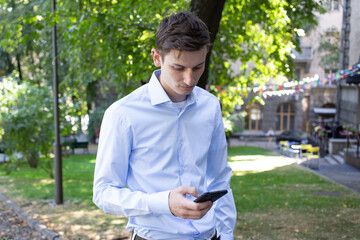 Portrait of young man in a blue shirt on the city with a phone in his hand looks at phone with a trees on the background. The man in conversation. American or European appearance. Summer day.