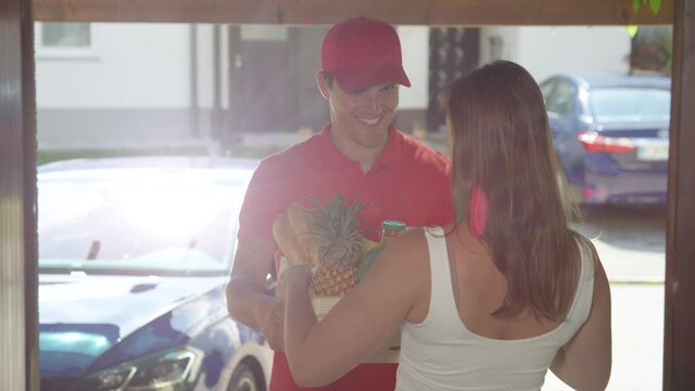 SLOW MOTION, LENS FLARE, CLOSE UP, DOF: Happy delivery guy hands Caucasian woman her order of organic produce. Smiling Caucasian courier wearing a red shirt and hat delivers groceries to a young woman