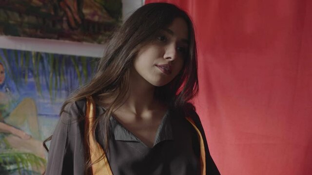 long haired girl ties ribbons of orange vest and dances near wall with different pictures in art studio closeup slow motion