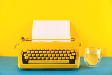 Yellow bright typewriter on a yellow and blue background next to glass of water. Symbol for writing, blogging, new ideas and creativity