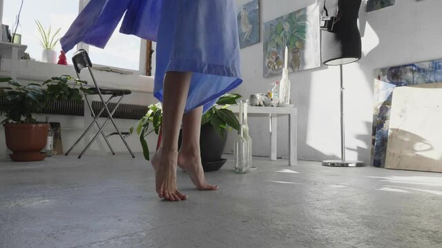 graceful young barefoot ballerina in long loose blue dress dances in modern art studio closeup slow motion
