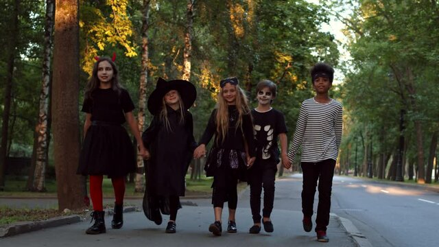 Wide shot of five cheerful multiethnic teenagers wearing Halloween costumes are going holding hands along city street