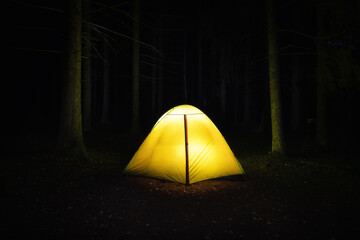 Camping in the forest. Orange illuminated tent under dark night trees. Illuminating tree trunks nearby.
