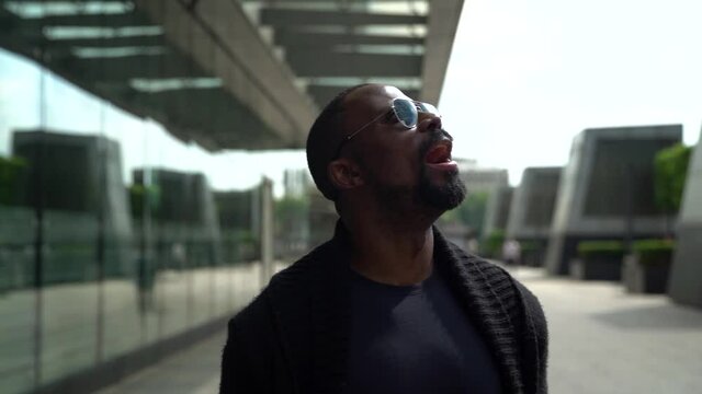 Portrait Of An African-American Man Walking Down A City Street During The Day And Looking Around At Buildings. He Wears Dark Glasses.