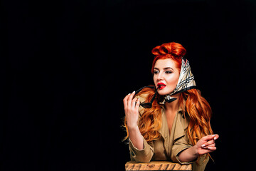A beautiful red-haired girl on a black background dressed in the style of pin-up with red lipstick holds glasses and does not look at the camera. Valentine's Day, March 8, International Women's Day.