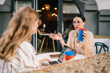 Positive female freelancers drinking coffee and discussing work in street cafe