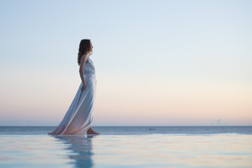 Fototapeta na wymiar Romantic photo of graceful woman walking in flowing dress along the water edge against gentle sunset sky