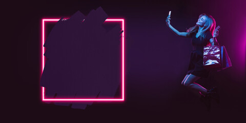 Selfie, jumping. Portrait of young woman in neon on dark studio backgound. Human emotions, black...