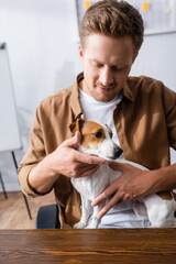 young businessman in casual clothes holding jack russell terrier dog while sitting at workplace