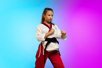 Practice. Karate, taekwondo girl with black belt isolated on gradient background in neon light. Little caucasian model, sport kid training in motion and action. Sport, movement, childhood concept.