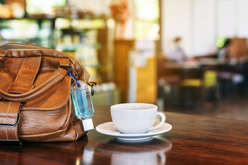 Mini alcohol gel bottle to kill Corona Virus(Covid-19) hang on a brown leather shoulder bag with hot coffee on table in cafe.New normal lifestyle.Health care concept.Selective focus on alcohol gel